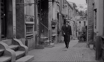 Movie still from “Le Doulos” (1962), directed by Jean-Pierre Melville – A black and white photo of a man walking down a street; Wide shot, Low angle