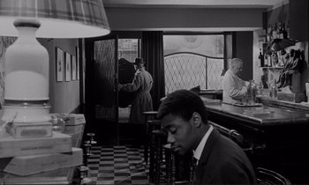 Movie still from “Le Doulos” (1962), directed by Jean-Pierre Melville – A man standing in a room next to a counter; Medium shot, High angle