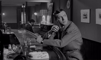 Movie still from “Le Doulos” (1962), directed by Jean-Pierre Melville – A black and white photo of a man sitting at a bar; Medium shot, Low angle