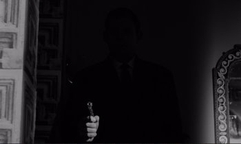 Movie still from “Le Doulos” (1962), directed by Jean-Pierre Melville – A man in a black suit holding a gun in the dark; Medium shot, Low angle