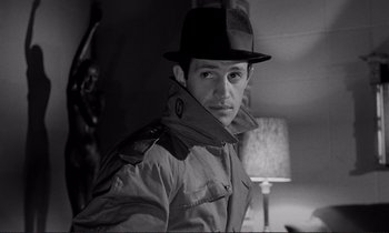 Movie still from “Le Doulos” (1962), directed by Jean-Pierre Melville – A man wearing a hat and a trench coat in front of a lamp; Close Up shot, Over the shoulder angle