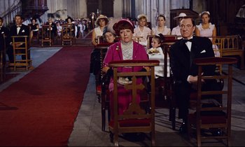 Movie still from “Le Grand Amour” (1969), directed by Pierre Étaix – An older woman sitting in a chair in front of a group of people; Wide shot, High angle