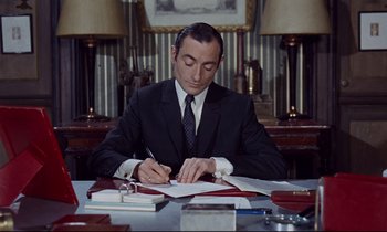 Movie still from “Le Grand Amour” (1969), directed by Pierre Étaix – A man sitting at a desk writing on a piece of paper; Medium shot, High angle