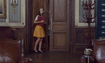 Movie still from “Le Grand Amour” (1969), directed by Pierre Étaix – A woman standing in front of a door holding a book; Wide shot, Over the shoulder angle