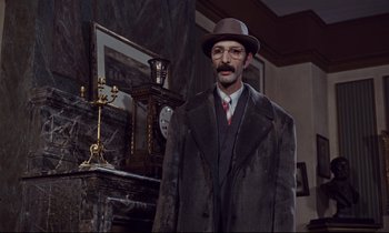Movie still from “Le Grand Amour” (1969), directed by Pierre Étaix – A man in a hat and a coat standing in front of a fireplace; Medium shot, Low angle