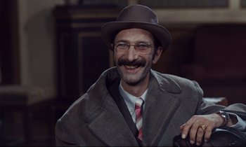 Movie still from “Le Grand Amour” (1969), directed by Pierre Étaix – A man in a hat and glasses smiling for the camera; Close Up shot, Over the shoulder angle
