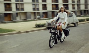 Movie still from “Le Grand Amour” (1969), directed by Pierre Étaix – A woman riding a bicycle down a street; Wide shot, High angle