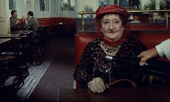 Movie still from “Le Grand Amour” (1969), directed by Pierre Étaix – An older woman sitting at a table holding a glass of wine; Medium shot, Over the shoulder angle