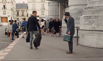 Movie still from “Le Grand Amour” (1969), directed by Pierre Étaix – A man in a suit and a hat talking on a cell phone while another man in a suit holds a guitar case in front of him; Wide shot, Low angle