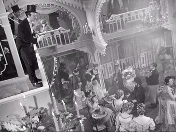 Movie still from “Le Plaisir” (1952), directed by Max Ophüls – A man in a top hat standing on a ladder in front of a group of people; Wide shot, High angle