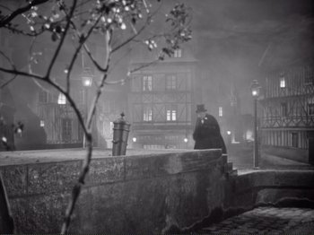 Movie still from “Le Plaisir” (1952), directed by Max Ophüls – A man sitting on a wall at night in front of a building; Extreme Wide shot, High angle