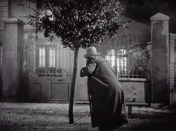 Movie still from “Le Plaisir” (1952), directed by Max Ophüls – A man in a hat and a long coat leaning against a tree in a park; Wide shot, Low angle