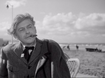 Movie still from “Le Plaisir” (1952), directed by Max Ophüls – An older man sitting on a chair smoking a cigarette; Medium shot, Low angle
