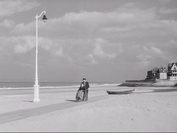 Movie still from “Le Plaisir” (1952), directed by Max Ophüls – A man riding a bike down a beach next to the ocean; Extreme Wide shot, High angle