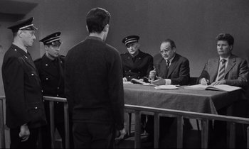 Movie still from “The Hole” (1960), directed by Jacques Becker – A group of men sitting at a table with a man standing in front of them; Medium shot, Over the shoulder angle
