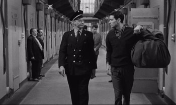 Movie still from “The Hole” (1960), directed by Jacques Becker – A man in a uniform walking down a hallway with another man; Medium shot, Over the shoulder angle