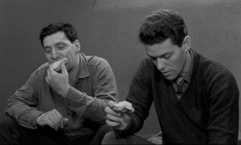 Movie still from “The Hole” (1960), directed by Jacques Becker – A couple of men sitting next to each other eating food; Medium shot, High angle