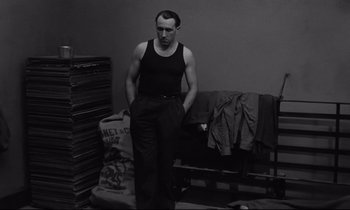 Movie still from “The Hole” (1960), directed by Jacques Becker – A man standing in front of a couch in a room; Medium shot, Low angle
