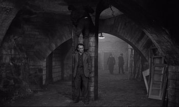 Movie still from “The Hole” (1960), directed by Jacques Becker – A man in a suit and tie standing next to a brick pillar; Wide shot, High angle