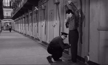 Movie still from “The Hole” (1960), directed by Jacques Becker – A man and a woman in uniform are standing in front of a wall; Wide shot, High angle