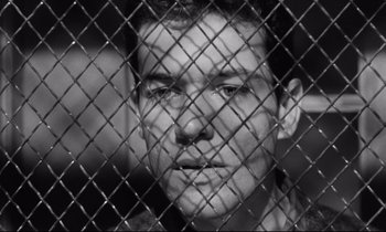 Movie still from “The Hole” (1960), directed by Jacques Becker – A black - and - white photo of a man behind a chain link fence; Close Up shot, Low angle