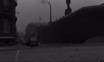 Movie still from “The Hole” (1960), directed by Jacques Becker – Cars parked on the side of the road near a factory; Extreme Wide shot, Low angle
