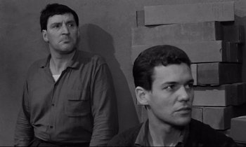 Movie still from “The Hole” (1960), directed by Jacques Becker – Two men are standing next to each other in front of a brick wall; Medium shot, Low angle