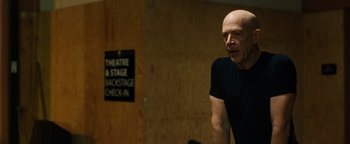 Movie still from “Whiplash” (2014), directed by Damien Chazelle – A bald man in a black t - shirt is standing in front of a sign; Medium shot, Low angle