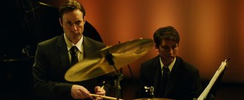 Movie still from “Whiplash” (2014), directed by Damien Chazelle – A man in a suit and tie playing drums; Medium shot, Over the shoulder angle