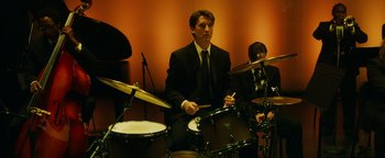 Movie still from “Whiplash” (2014), directed by Damien Chazelle – A man in a suit and tie playing drums in front of other men; Medium shot, Low angle