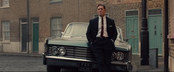 Movie still from “Legend” (2015), directed by Brian Helgeland – A man standing next to a car in front of a brick building; Medium shot, Low angle