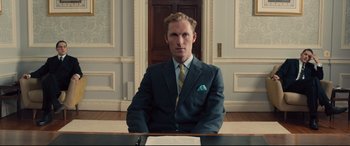 Movie still from “Legend” (2015), directed by Brian Helgeland – A man sitting at a desk in a suit and tie; Medium shot, Low angle