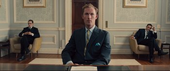 Movie still from “Legend” (2015), directed by Brian Helgeland – A man sitting at a desk in a suit and tie; Medium shot, Low angle