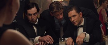 Movie still from “Legend” (2015), directed by Brian Helgeland – A group of men in suits sitting at a table with drinks; Medium shot, Low angle