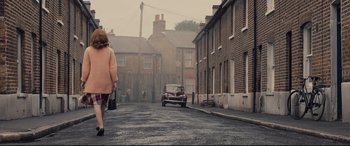 Movie still from “Legend” (2015), directed by Brian Helgeland – An older woman walking down a street with a car; Wide shot, High angle