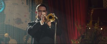 Movie still from “Legend” (2015), directed by Brian Helgeland – A man in a black suit playing a trombone; Medium shot, Over the shoulder angle