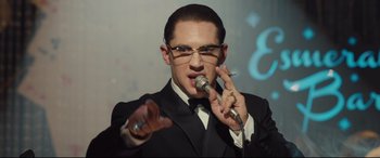 Movie still from “Legend” (2015), directed by Brian Helgeland – A man in a black suit is singing into a microphone; Close Up shot, Over the shoulder angle