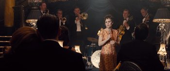 Movie still from “Legend” (2015), directed by Brian Helgeland – A woman holding a microphone in front of a group of people; Medium shot, Over the shoulder angle