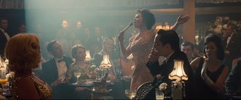 Movie still from “Legend” (2015), directed by Brian Helgeland – A woman singing into a microphone in front of a group of people; Medium shot, Over the shoulder angle