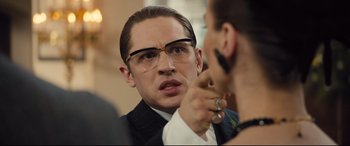 Movie still from “Legend” (2015), directed by Brian Helgeland – A man in a suit and glasses talking to a woman in a dress suit; Close Up shot, Over the shoulder angle