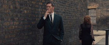 Movie still from “Legend” (2015), directed by Brian Helgeland – A man in a suit smoking a cigarette in front of a brick wall; Medium shot, Low angle