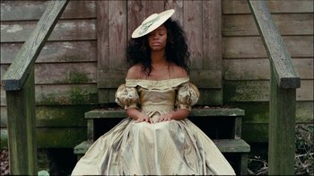 Movie still from “Lemonade” (2016), directed by Mark Romanek – A woman wearing a dress and a hat sitting on top of a wooden bench; Medium shot, High angle