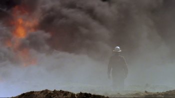 Movie still from “Lessons of Darkness” (1992), directed by Werner Herzog – A man in a white hat standing in front of a smoke filled sky; Extreme Wide shot, Low angle
