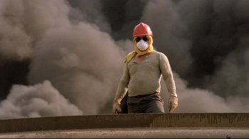 Movie still from “Lessons of Darkness” (1992), directed by Werner Herzog – A man wearing a hard hat and a gas mask while standing in front of a smoke filled sky; Medium shot, Low angle