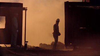 Movie still from “Lessons of Darkness” (1992), directed by Werner Herzog – A man in a hard hat walking on a dirt road; Wide shot, Low angle