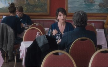 Movie still from “Let the Sunshine In” (2017), directed by Claire Denis – A woman sitting at a table holding a glass of wine; Medium shot, Over the shoulder angle
