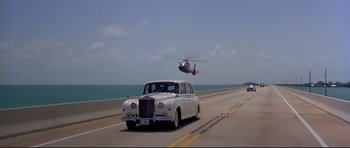 Movie still from “Licence to Kill” (1989), directed by John Glen – An old car driving down a highway with a helicopter in the background; Extreme Wide shot, High angle