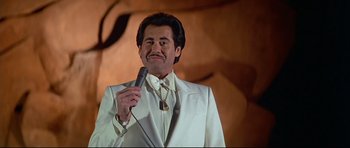 Movie still from “Licence to Kill” (1989), directed by John Glen – A man in a white suit is holding a microphone; Medium shot, Low angle