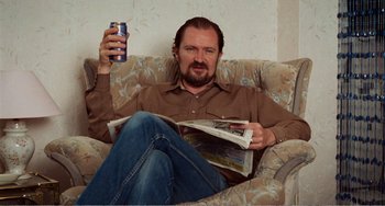 Movie still from “Life Is Sweet” (1990), directed by Mike Leigh – A man sitting on a couch holding a drink and reading a newspaper; Medium shot, High angle