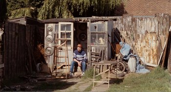 Movie still from “Life Is Sweet” (1990), directed by Mike Leigh – A man sitting in front of an outhouse; Wide shot, High angle